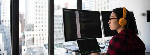 Mujer programando en frente de una ventana, utilizando varias pantallas.